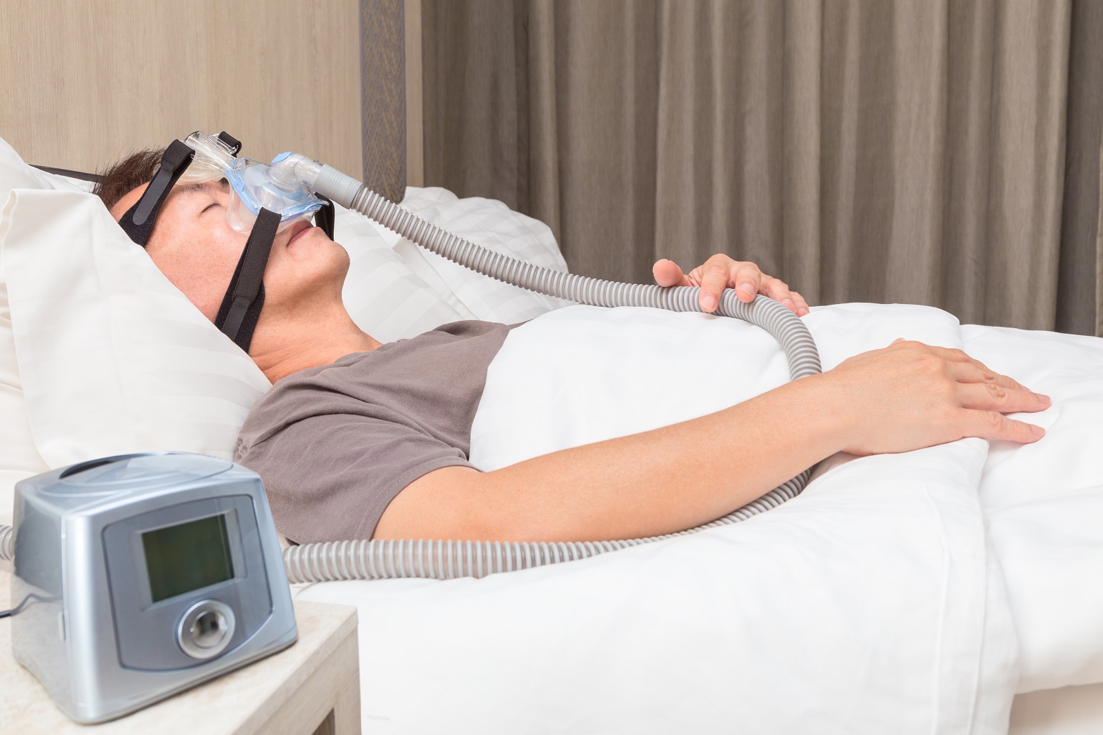 Middle age Asian man sleeping in his bed wearing CPAP mask connecting to air hose and CPAP machine